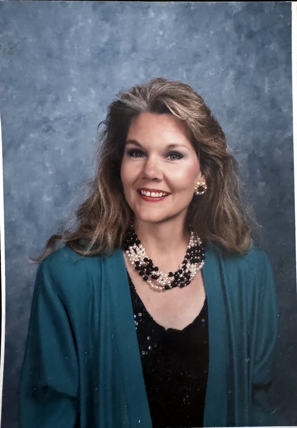 Studio portrait of Eve Hess smiling, in a teal jacket and a pearl and black bead necklace